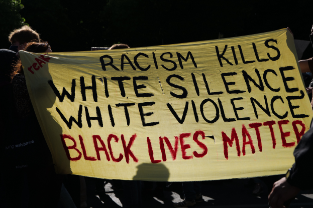 Eine Gruppe von Menschen steht auf einer Straße und hält ein Banner mit der Aufschrift "Rassismus tötet Weißes Schweigen, weiße Gewalt, Black Lives Matter", mit Bäumen im Hintergrund.