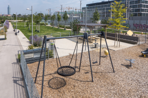 Ein Spielplatz mit Schaukelgerüst umgeben von Bäumen, Pflanzen, Gras, Straßenmasten, Straßenlaternen, Strommasten, Stromkabeln, Gebäuden und einem bewölkten Himmel.