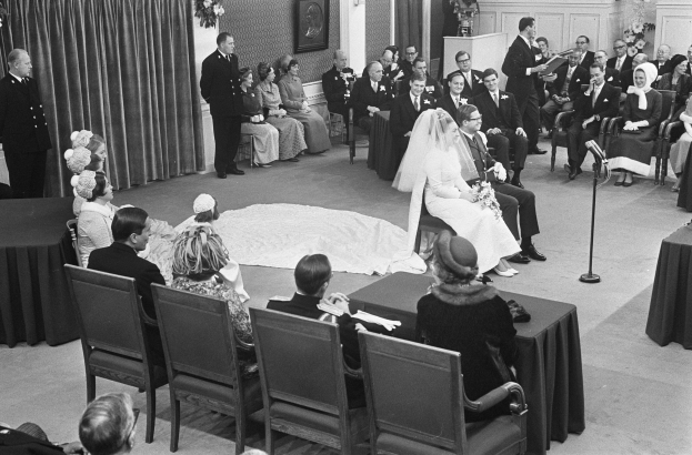 Schwarzes und weißes Foto einer Hochzeitszeremonie mit sitzenden und stehenden Gästen, einem Mann, der in ein Mikrofon spricht, einer Frau mit einem Blumenstrauß, einem Podium mit einem Mikrofon, Blumengestecken, einem Vorhang, einem gerahmten Foto an der Wand und einer Tür im Hintergrund.