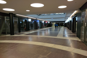 Langer Flur mit Menschen, die gehen, beleuchtet von Deckenlampen, gesäumt von Säulen, Glastüren und Texttafeln, ähnelt einem belebten Rathaus.