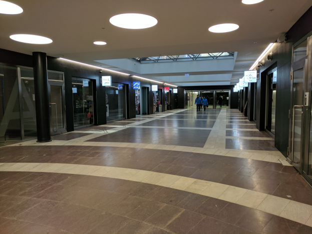 Langer Flur mit Menschen, die gehen, beleuchtet von Deckenlampen, gesäumt von Säulen, Glastüren und Texttafeln, ähnelt einem belebten Rathaus.