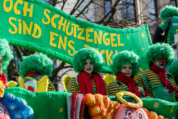 Eine Gruppe von Clowns in bunten Kostümen marschiert durch eine Straße und hält ein Banner mit Text, mit Bäumen und einem Gebäude im Hintergrund.