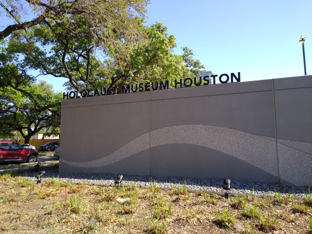 Außenansicht des Houston Holocaust Museums mit einer Textwand, Gedenksteine, Landschaft, Straßenverkehr und einem bewölkten Himmel.