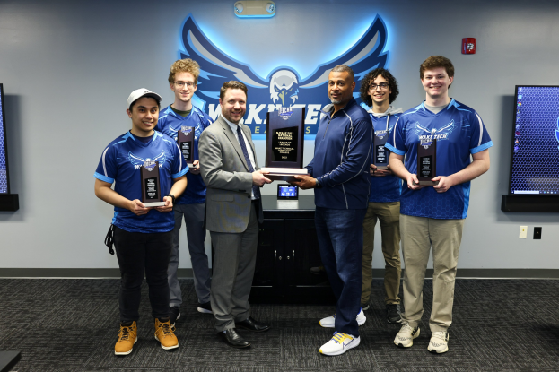 Gruppe von lachenden Männern mit Trophäen, die vor zwei Fernsehern mit Logo und Text an der Wand dahinter stehen.