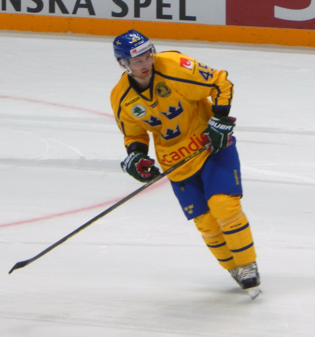 Eishockeyspieler in Bewegung auf dem Eis mit Helm, Handschuhen, Schuhen und einem Hockeystock in der Hand, mit einer Tafel im Hintergrund, auf der Text steht.