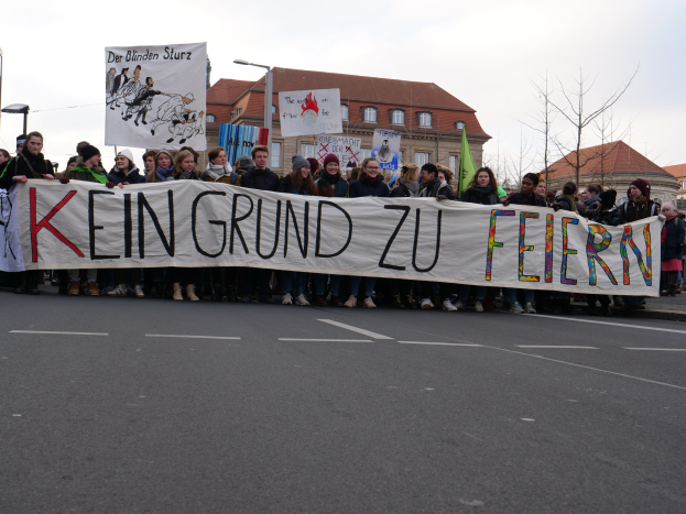 Demonstranten mit einem Transparent mit der Aufschrift "Kein Grund zu Feiern" stehen auf einer Straße mit Laternenmasten, Straßenlaternen, Bäumen und Gebäuden im Hintergrund bei klarem blauem Himmel.