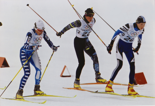 Drei Skifahrer, die eine schneebedeckte Piste hinunterrasen, jeder hält Ski-Stöcke in der Hand, trägt bunte Ski-Anzüge und Helme, der Schnee glitzert in der Sonne.