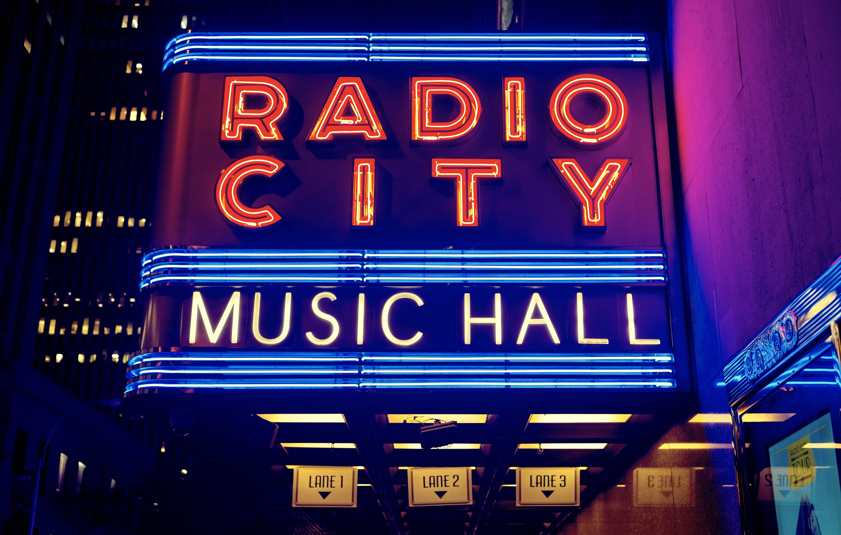 Radio City Music Hall in New York City, hell erleuchtet und von hohen Gebäuden flankiert, mit einer 'Radio City'-Leuchtschrift und zusätzlichen Schildern auf der rechten Seite.