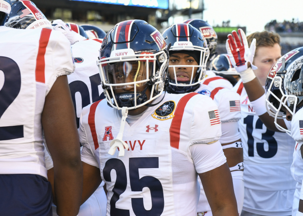 Gruppe von Männern in Football-Uniformen auf einem Feld mit Helmen und Handschuhen, Hintergrund zeigt eine unscharfe Menge, ein Scoreboard mit der Aufschrift "Auburn vs Virginia Tech - 11/28/15 College Football Pick, Odds, and Prediction" und Himmel.