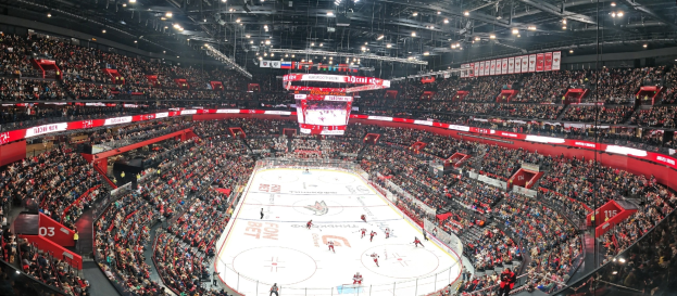 Eine große, hell erleuchtete Arena mit Zuschauern, die ein Hockey-Spiel verfolgen, mit einem zentralen Bildschirm über der Eisfläche und Fans, die in den Rängen jubeln.