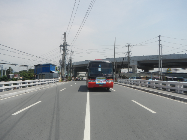 Ein roter Bus fährt auf einer Autobahn mit weißen Linien, umgeben von Leitplanken, mit Strommasten, einer Brücke, Bäumen, Gebäuden und einem klaren blauen Himmel im Hintergrund.
