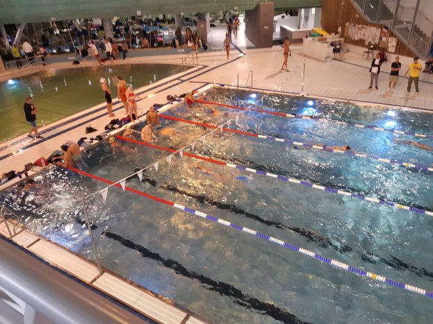 Großes Hallenbad mit schwimmenden, stehenden und sitzenden Menschen, einschließlich Treppe und Geländer auf der rechten Seite.
