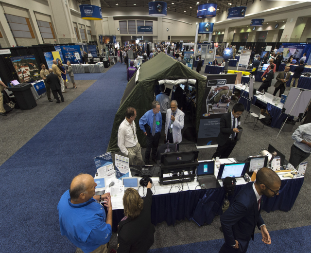 Eine große Gruppe von Menschen auf einer Karriere-Messe um einen Tisch mit Laptops, Monitoren und Papieren versammelt, mit einem Zelt, Schautafeln, Deckenleuchten und Fenstern im Hintergrund.