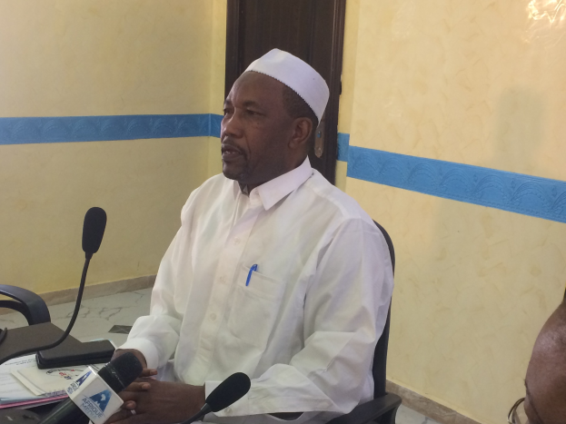 Ein Mann in einem weißen Kleid und einer Mütze sitzt an einem Tisch mit Mikrofonen, spricht, während Papiere und Gegenstände auf dem Tisch liegen, und ein weiterer Mensch mit Brille rechts von ihm in einem Raum mit einer Wand und einer Tür im Hintergrund.