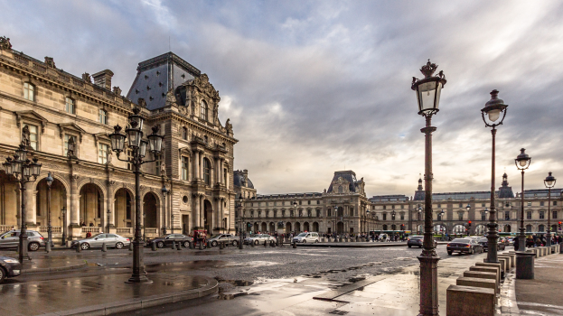 Außenansicht des Louvre-Museums in Paris, Frankreich, mit seiner ikonischen Architektur, Straßenlaternen, fahrenden Fahrzeugen, Fußgängern auf dem Gehweg und einer bewölkten Himmel.