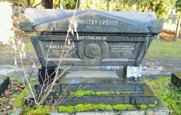 Ein Grabstein mit Moosbewuchs in einem Friedhof, mit Bäumen im Hintergrund.