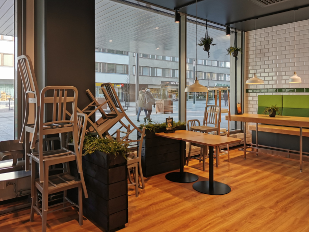 Ein Restaurant-Innenraum mit hölzernen Tischen und Stühlen, großen Fenstern, Pflanzen und Beleuchtung, mit Blick auf eine Berliner Straße mit Fußgängern, Gebäuden und urbanen Elementen.