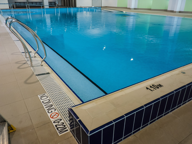 Großes Hallenbad in einem großen Gebäude mit gefliestem Boden, Metallträgern, einer Schautafel, einer Tür, einer Wand und Deckenleuchten.