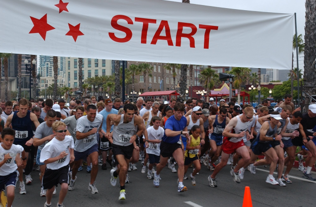 Gruppe von Menschen beim Marathonlauf mit einem Verkehrskegel im Vordergrund und einem Banner mit Text im Hintergrund, umgeben von Bäumen, Laternenmasten, Gebäuden und einem klaren blauen Himmel.