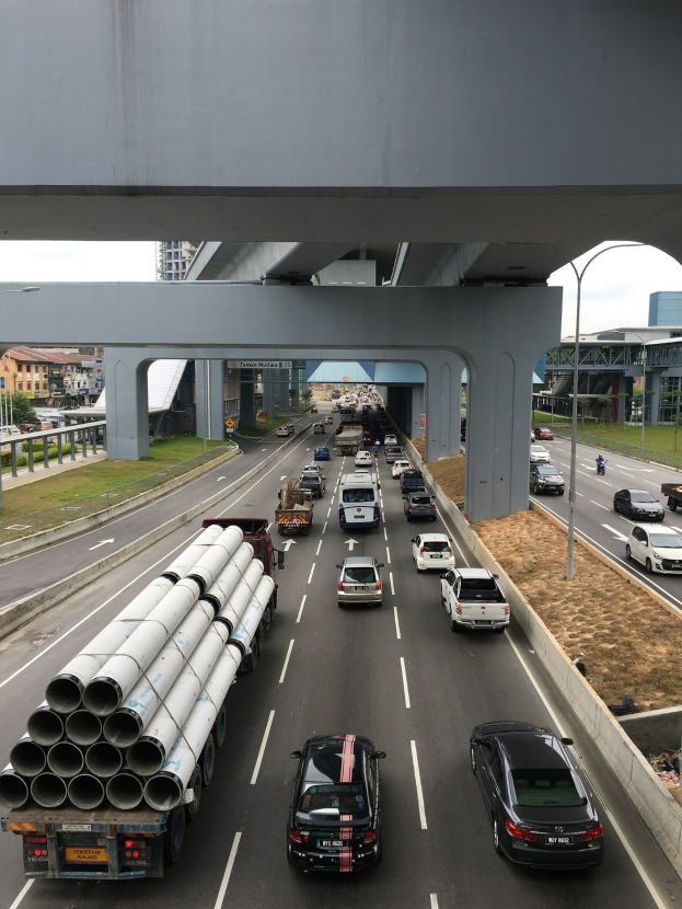 Eine vielbefahrene Autobahn mit starkem Verkehr unter einer Brücke, umgeben von Gras, Bäumen, Gebäuden und Strommasten.