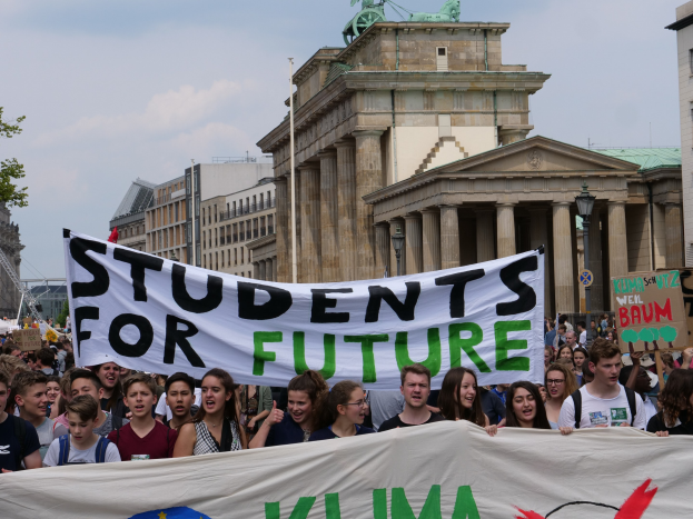 Eine Gruppe von Studenten marschiert in Berlin mit einer bunt bemalten "Students for Future"-Schlagzeile gegen Gebäude, Bäume und Himmel.
