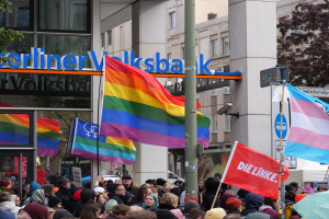 Eine große Gruppe von Menschen mit Fahnen und Schildern steht vor einem Gebäude, mit einem Mast im Vordergrund und Bäumen zu beiden Seiten, bei einer Gay-Pride-Parade in Berlin.