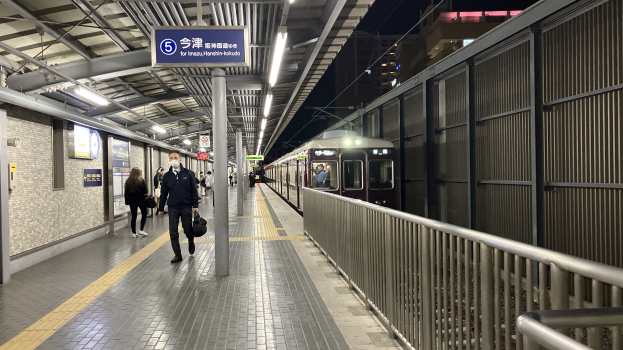 Ein Zug, der auf einem Bahnsteig einfährt, mit Menschen in der Nähe, einem Geländer auf der rechten Seite, Gebäuden im Hintergrund, Bahnsteigbeleuchtung und einer Anzeigetafel.