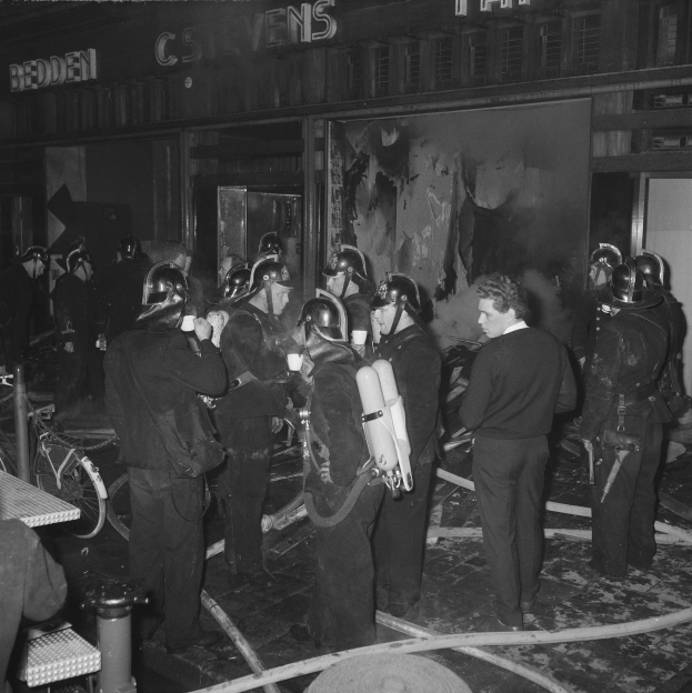 Schwarzes Bild von Feuerwehrleuten in Uniform mit Atemschutz, die vor einem Gebäude, einem Fahrrad und einem Tisch mit Gegenständen stehen.