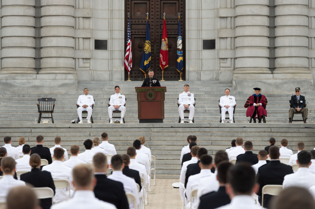 Gruppe von Menschen, die auf Stühlen vor einem Gebäude mit Säulen und einer Tür sitzen, mit einem Podium und einem Mikrofon in der Mitte, Flaggen im Hintergrund und einem leeren Stuhl links, was auf eine Abschlussfeier hinweist.