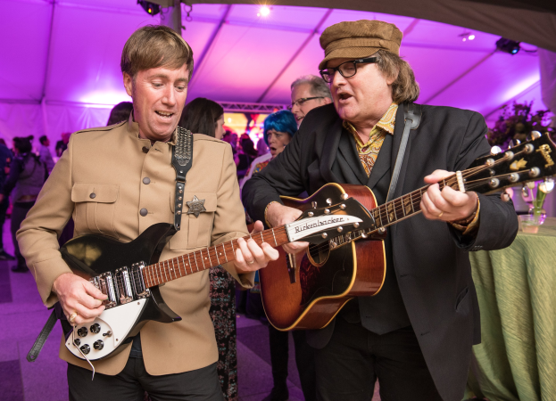 Zwei Männer, die in einem Zelt Gitarre spielen, umgeben von einer Menge, ein grüner Tisch mit Gläsern und einer Vase auf der rechten Seite und ein beleuchtetes Zeltdach darüber.