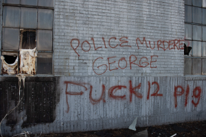 Ein Gebäude mit Graffiti, das "Polizei Mord George Fuck 12 P19" auf der Seite trägt, mit Fenstern und verschiedenen Gegenständen vorne.