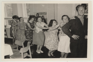 Eine Gruppe von Menschen, die in einem Raum im Stil der 1950er Jahre tanzen, mit Stühlen, einem Tisch, einem Foto Rahmen an der Wand und Fenstern im Hintergrund.