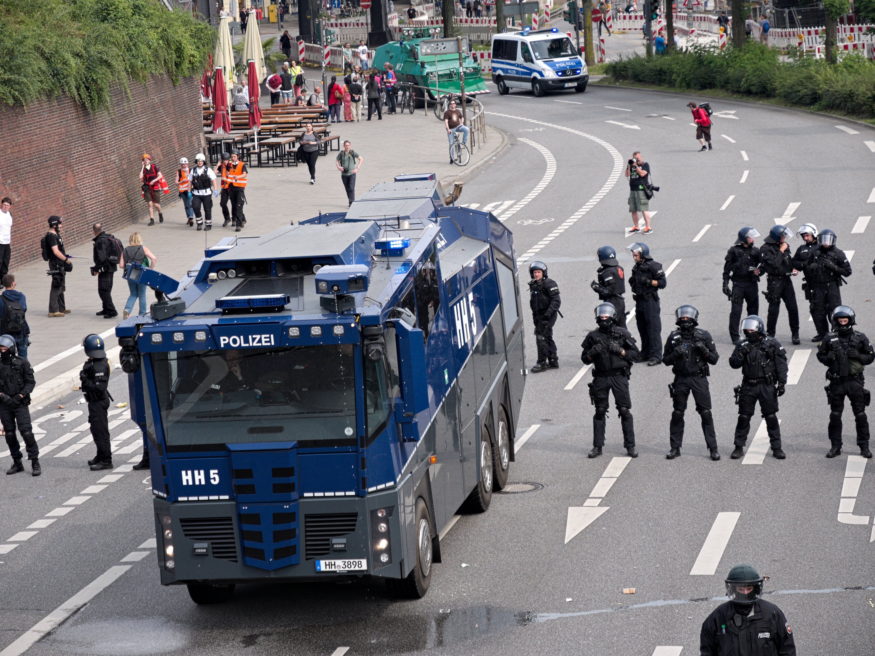 Gruppe von Polizisten vor einem gepanzerten Fahrzeug auf einer Stra├če mit bewaffneten Personen, umgeben von urbanen Elementen wie Geb├Ąuden, Verkehrszeichen und Vegetation.
