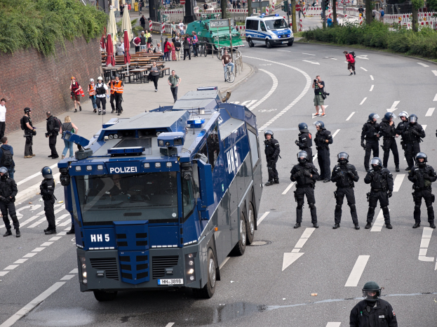 Gruppe von Polizisten vor einem gepanzerten Fahrzeug auf einer Stra├če mit bewaffneten Personen, umgeben von urbanen Elementen wie Geb├Ąuden, Verkehrszeichen und Vegetation.