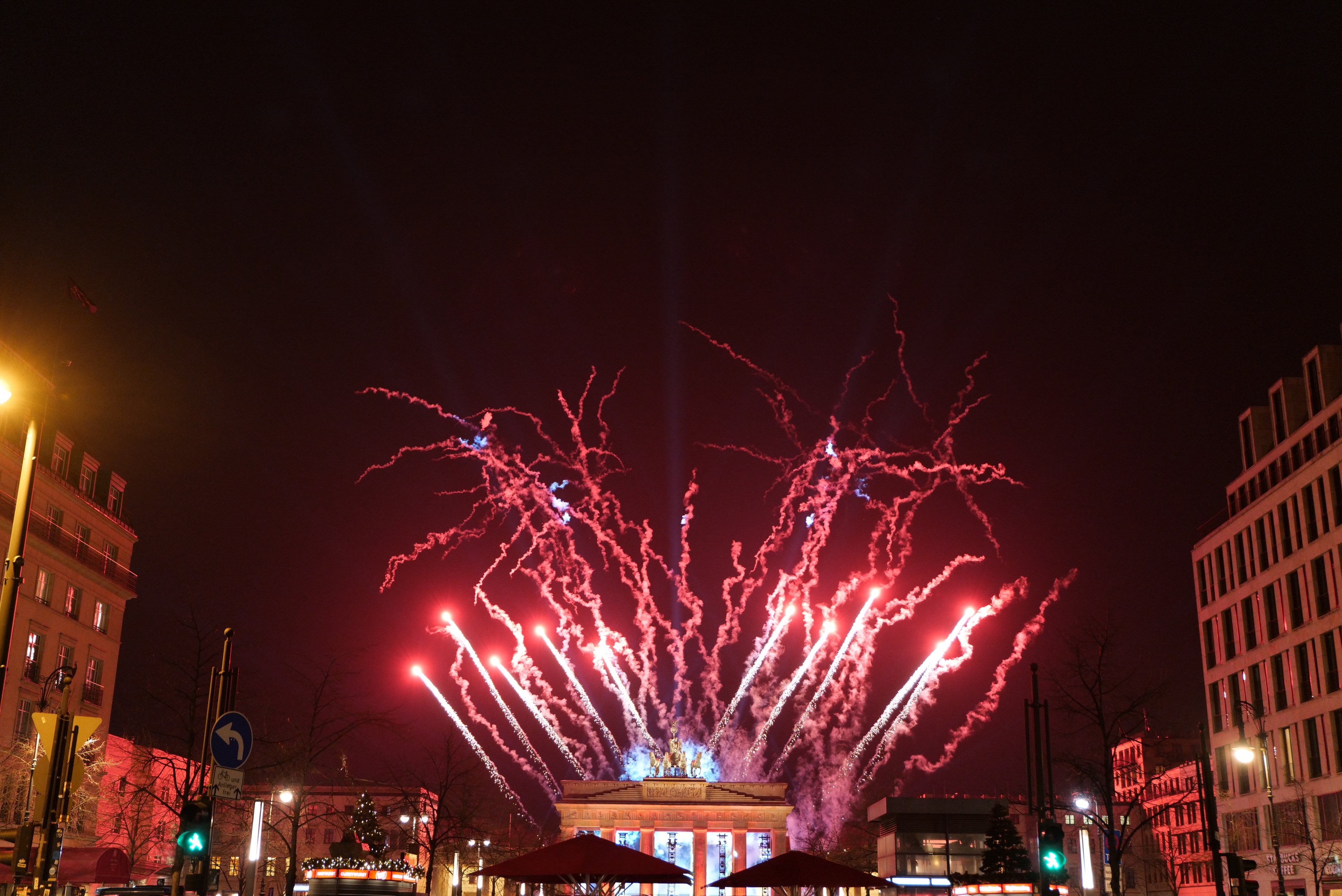 Eine belebte Stadtstraße an Silvester in Berlin, voller Menschen, Fahrzeuge und festlicher Dekoration, mit Feuerwerk, das den Himmel und die Gebäude erhellt.