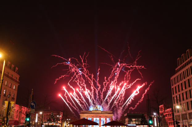 Eine belebte Stadtstraße in Berlin am Silvesterabend, voller Menschen, Fahrzeuge und festlicher Beleuchtung, mit Feuerwerk, das den Himmel über den Gebäuden erhellt.