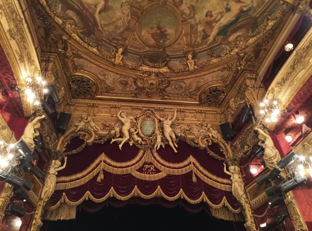 Theaterinnenraum mit roten Vorhängen, Kronleuchtern, Deckenmalerei und Skulpturen, mit einer Bühne mit ein paar Menschen davor.