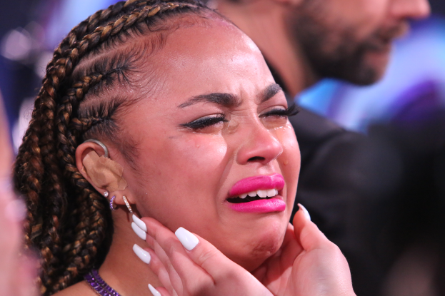 Eine Frau mit Zöpfen und pinkem Lippenstift weint im Vordergrund, mit unscharfen Menschen und Hintergrund dahinter.