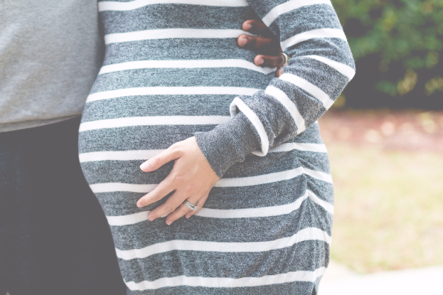 Schwangeres Paar, das Hand in Hand mit Händen auf dem Bauch steht, trägt Schwangerschaftskleidung in einem bewaldeten Outdoor-Gebiet.