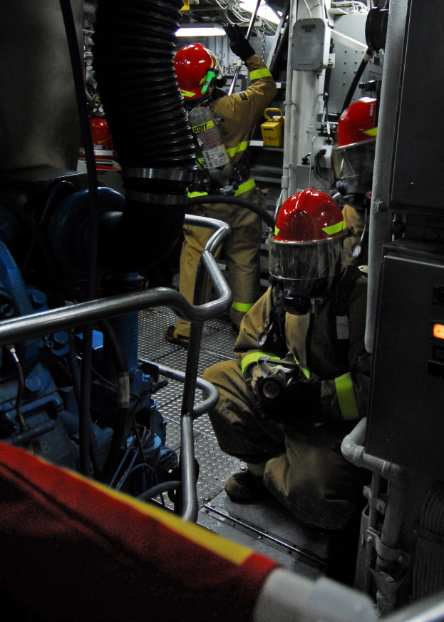 Feuerwehrleute in Helmen und Schutzausrüstung arbeiten an einem Feuerlöschfahrzeug mit Rohren und einer Lampe im Hintergrund.