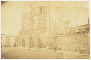 Altes Foto eines großen Gebäudes in einer städtischen Umgebung, umgeben von kleineren Gebäuden, Straßenlaternen, Straßenlaternen und einer Straße.