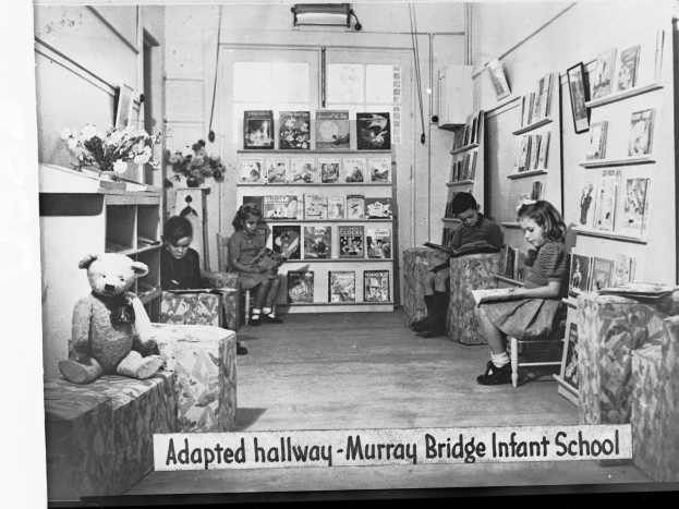 Eine Gruppe von Kindern sitzt auf Stühlen in einem Raum mit Regalen, einem Teddybär auf der linken Seite, einer Blumenvase auf dem Tisch und gerahmten Gegenständen an der Wand, mit dem Text "Adapted Hallway Murray Bridge Infant School" unten.