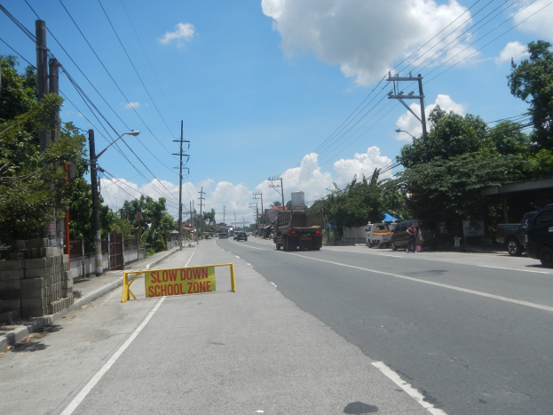 Straße mit Fahrzeugen, ein "Slow Down School Zone"-Schild, Strommäste mit Drähten, Bäume, Laternenpfähle und ein bewölkter Himmel im Hintergrund.