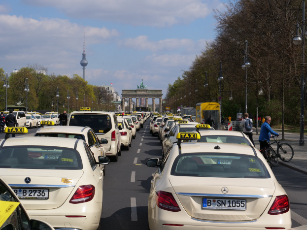 Eine lange Reihe von Taxis, die entlang einer belebten Straße in Berlin, Deutschland, geparkt sind, mit Fahrzeugen, Radfahrern und Fußgängern; Laternenpfähle und Bäume säumen den Gehweg, und Gebäude, ein Bogen und ein Turm stehen im Hintergrund.