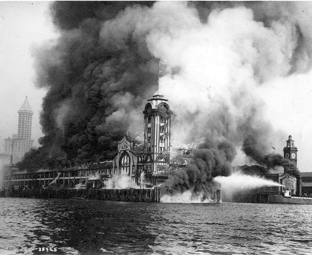 Schwarzes und weißes Foto eines großen, von Flammen umloderten Gebäudes mit Rauchwolken, einem Gewässer im Vordergrund und anderen Gebäuden sowie dem Himmel im Hintergrund.