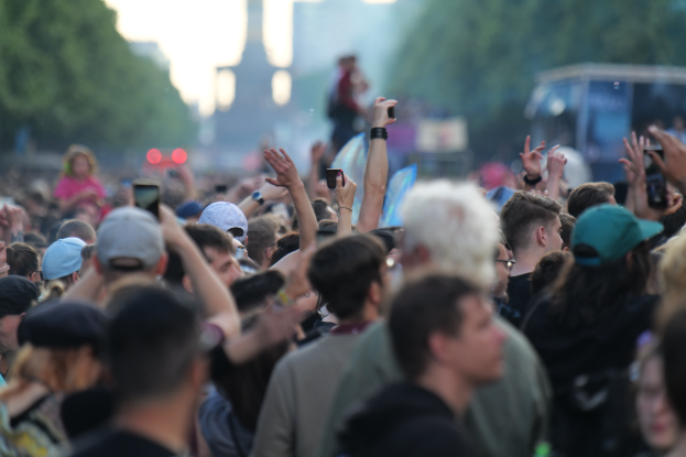 Eine Menschenmenge auf einem Musikfestival mit erhobenen Händen, einige tragen Mützen und halten Mobiltelefone, im Hintergrund sind Bäume, Gebäude, Lichter und ein Fahrzeug zu sehen.