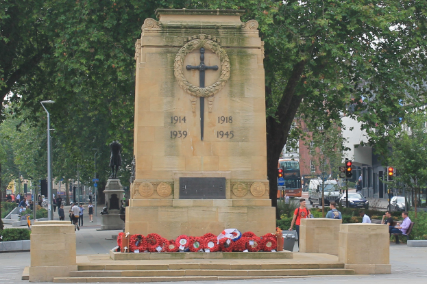 Kriegsdenkmal in einer Stadtstraße umgeben von Kränzen, mit Menschen auf Bänken, Fahrzeugen, Verkehrsampeln, Laternenpfählen, Bäumen, Gebäuden und Himmel im Hintergrund.