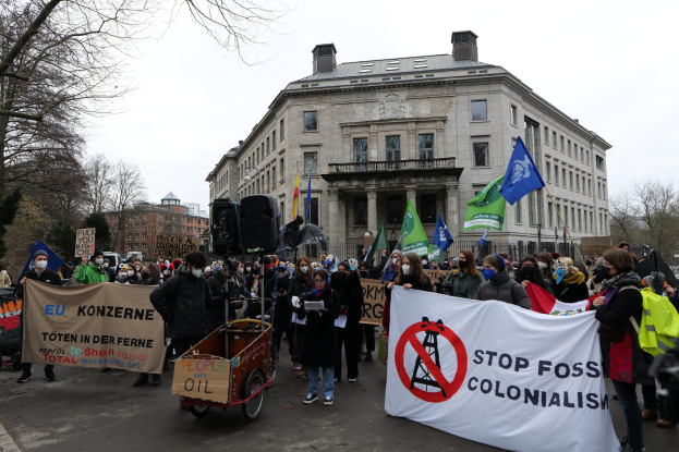 Eine große Gruppe von Menschen marschiert in einem Protest gegen fossile Brennstoffe, trägt Schilder und Fahnen und hat ein Fahrzeug im Vordergrund und Gebäude, Bäume und einen klaren blauen Himmel im Hintergrund.