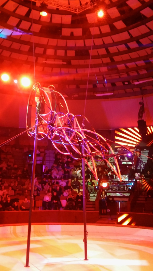 Ein Zirkusinneres mit einem großen Metallgerüst in der Mitte, umgeben von Stangen und Lichtern, mit Menschen im Hintergrund und diversen Gegenständen, die herumliegen, unter einer Decke mit Einbauleuchten.