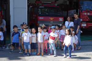 Eine Gruppe von Kindern vor einem Feuerwehrauto auf einer Feuerwache stehend, einige mit Mützen, sowie weitere Feuerwehrautos im Hintergrund.